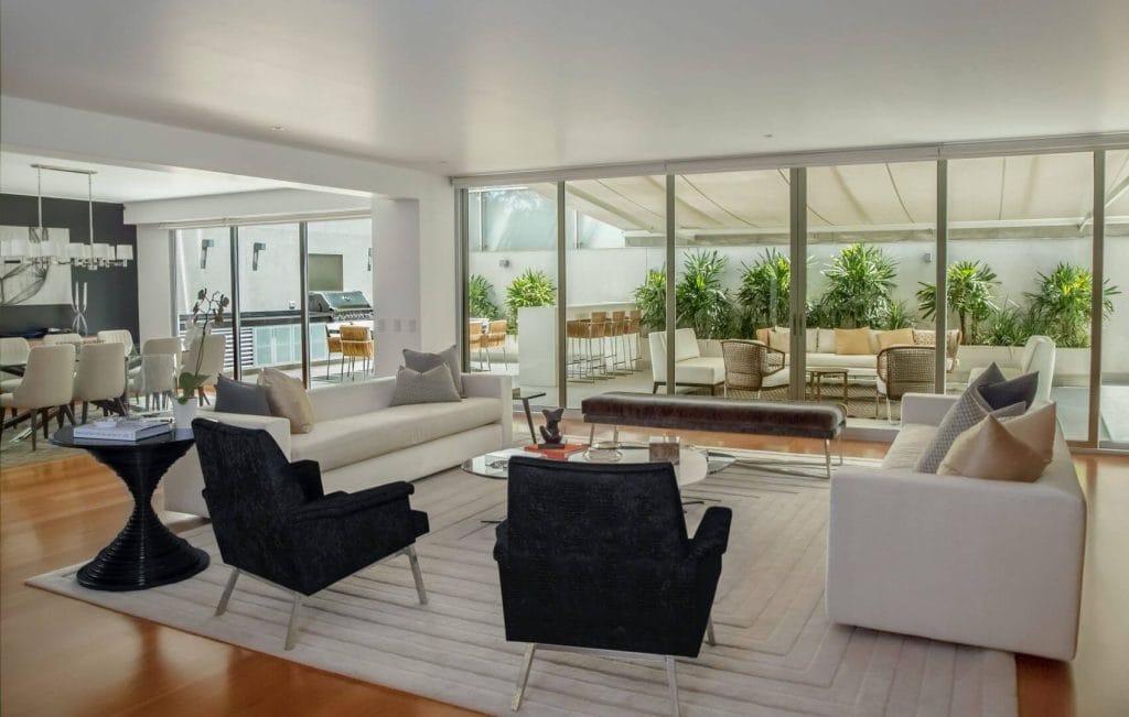 Spacious modern living room with white sofas, black armchairs, glass coffee table, and large sliding glass doors opening to an outdoor patio with seating and greenery.