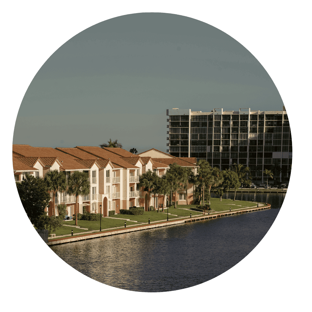 Row of waterfront condominiums with terracotta roofs, palm trees along a canal, and a larger residential building in the background under a clear sky.
