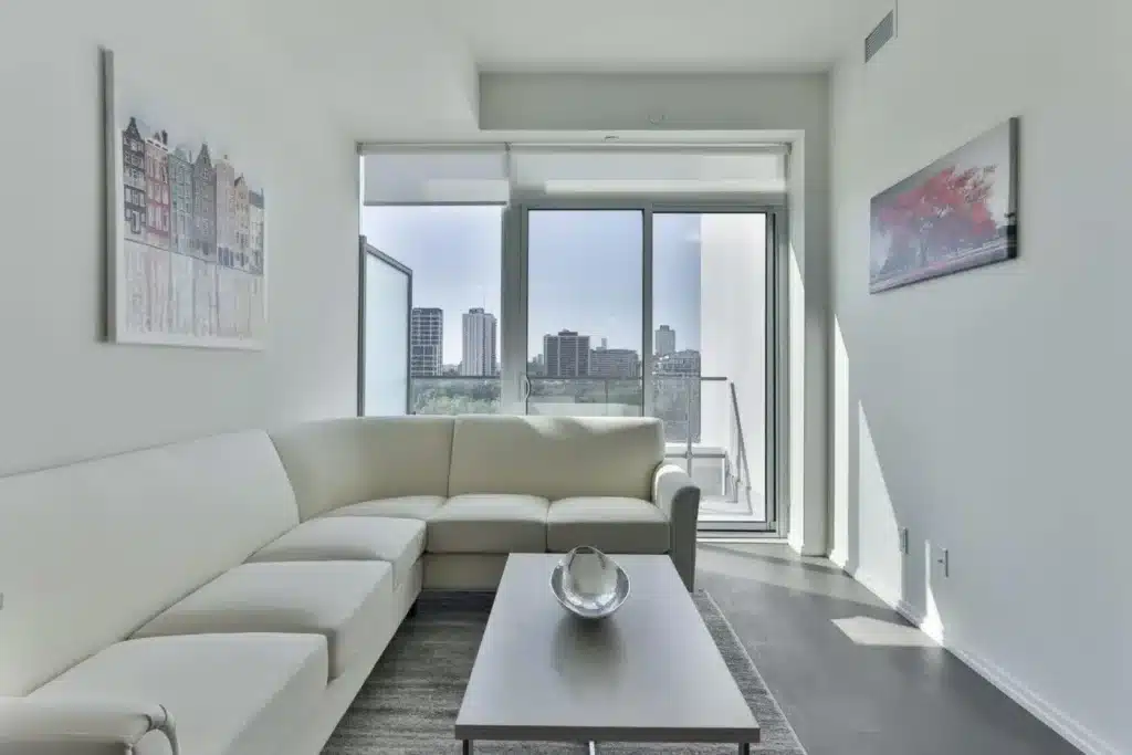 Modern living room with white sectional sofa, glass-top coffee table, large floor-to-ceiling windows, and city skyline view from a balcony.